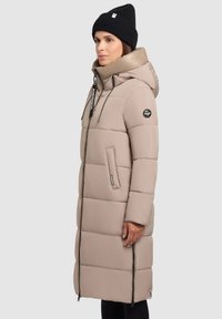 Pikk beez puffer-kate koos kapuutsi, millel on tikitud osad, küljetaskud ja lukku, täiendab must kudumüts.