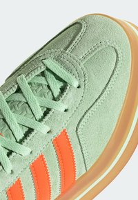 Zelené semišové tenisky s texturovaným povrchem, které mají oranžové zoubkované pruhy a odpovídající tkaničky, a nízkoprofilovou tan gumovou podrážku.