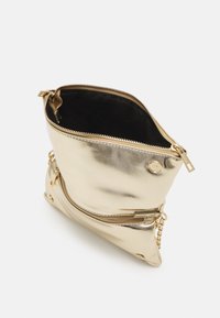 Petit sac bandoulière métallique doré avec fermeture éclair ouverte sur le dessus, poche avant zippée et bandoulière chaîne dorée sur fond blanc.