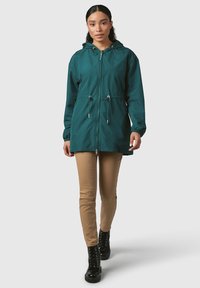 Giacca impermeabile turchese con cappuccio regolabile, chiusura con zip, polsini elasticizzati e vita regolabile. Indossata sopra pantaloni beige e stivali neri con lacci.