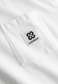 Witte stof met een gestikte zak waarop een zwart verweven logo en de tekst "GABBA" op een rechthoekig wit etiket zijn aangebracht.