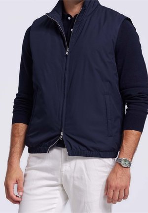 Mand iført navy blå lynlåsløs vest over sort langærmet skjorte med hvide bukser og sølvfarvet armbåndsur, hænder afslappet langs siderne.