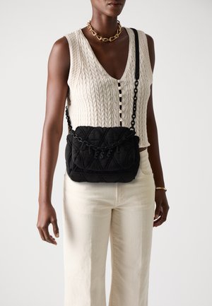 Person wearing a cream sleeveless knit top, cream pants, gold chain necklace, and black quilted crossbody bag with "DIESEL" letters.