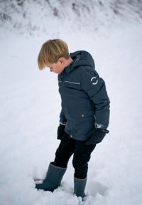 Otrok v temno modri zimski jakni z belimi poudarki, črnih hlačah in sivih škornjih, stoji v snegu, gleda navzdol.