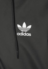 Černá mikina Adidas s texturovaným povrchem, s výrazným bílým logem trefoil a černými šňůrkami kapuce.