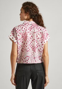 Camisa corta de manga corta en blanco con patrones geométricos y florales en rosa, con cuello puntiagudo y corte relajado.
