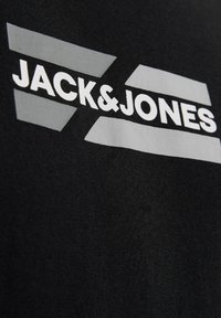 Λευκό κείμενο "JACK & JONES" με γκρίζα γεωμετρικά σχήματα σε μαύρο ύφασμα, πιθανώς τυπωμένο λογότυπο σε ρούχο.