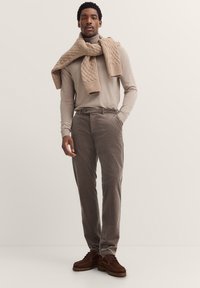 Beige gestrickter Schal, hellbeige Langarmshirt, graue Cordhosen und braune Schnürschuhe. Modell steht mit den Händen in den Taschen.