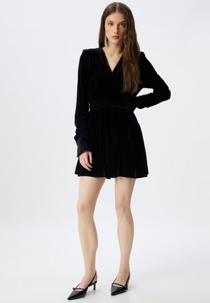 Vestito di velluto nero con design avvolgente, maniche lunghe, vita arricciata, gonna svasata e maniche lunghe. Indossato con tacchi a punta neri.