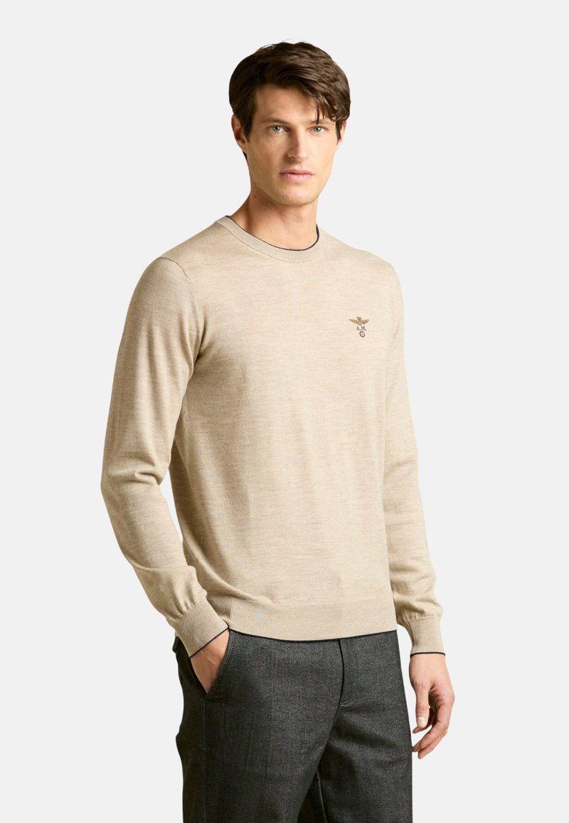Maglione beige a maniche lunghe con scollo rotondo e polsini a coste, caratterizzato da un piccolo logo ricamato sul petto sinistro. Tessuto leggero e strutturato.