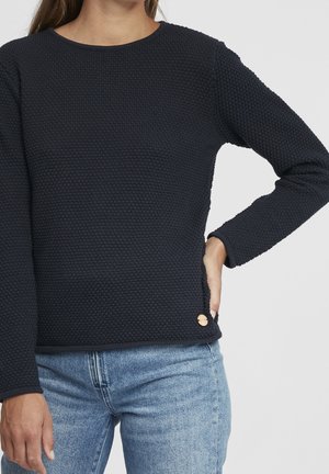 Frau trägt ein strukturiertes dunkelblaues Langarmshirt und hellblaue Jeans, Hand in der Hüfte, vor einem schlichten grauen Hintergrund.