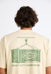 Beige short-sleeve t-shirt featuring a green graphic illustration of a shipping container, crane, and text "Dickies HEAVY DUTY WORKWEAR."