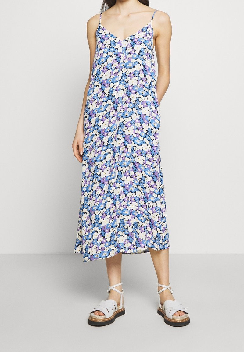 Kwiecista sukienka midi w niebieskie, fioletowe i białe kwiaty, dekolt w serek, cienkie ramiączka, zwiewna tkanina, w połączeniu z szarymi sandałami na paski.