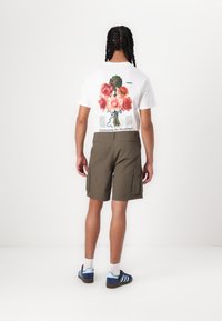 T-shirt blanc avec un graphisme floral coloré au dos, associé à des shorts cargo vert olive et des chaussures de sport avec des accents bleus.