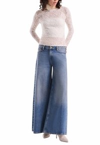 Weißes Spitzenoberteil mit langen Ärmeln, kombiniert mit weiten blauen Jeans. Die Jeans zeigen einen Farbverlauf von hell nach dunkel, haben eine glatte Textur und sichtbare Nähte.