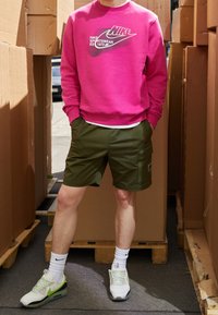 Růžová mikina s logem Nike, zelené cargo šortky s bočními kapsami a bílé tenisky se zelenými akcenty. Stojí mezi kartonovými krabicemi.