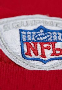 Fond en tissu rouge avec un gros plan sur un logo NFL brodé. Le patch présente un bouclier blanc avec des accents bleus et des lettres rouges.
