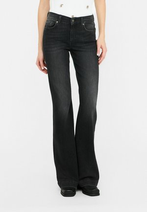 Kobieta w czarnych dzwonach z wysokim stanem, do których ma czarne buty i biały top z złotymi guzikami.