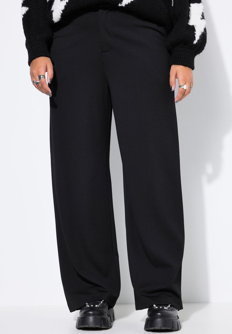 Schwarze weit geschnittene Hosen aus einem glatten Stoff, mit hoher Taille und einem klaren Design. Getragen mit klobigen schwarzen Schuhen.