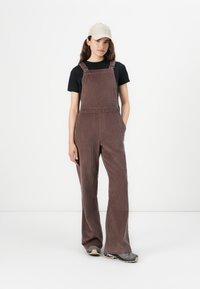 Bretelle in corduroy marrone con gambe larghi, spalline regolabili e tasche frontali, abbinate a una maglietta nera e un cappellino beige.