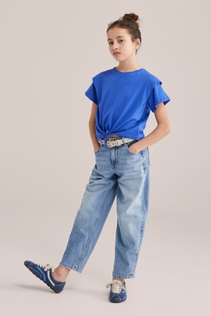 Kontyba kötött hajjal rendelkező lány kék rövid ujjú felsőben, amelyet deréknál megkötött, világoskék farmernadrágban és kék tornacipőben, kezét a zsebében tartva áll.
