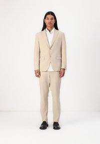 Giacca di abito beige con pantaloni abbinati, caratterizzata da un design a un bottone, due tasche frontali e scarpe nere. Camicia bianca sotto.