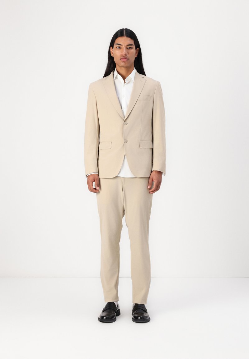 Giacca di abito beige con pantaloni abbinati, caratterizzata da un design a un bottone, due tasche frontali e scarpe nere. Camicia bianca sotto.