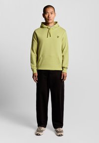 Ljusgrön hoodie med en framficka och dragsko på huvan, med en liten logotyp på bröstet. Kombinerad med svarta byxor och sneakers.