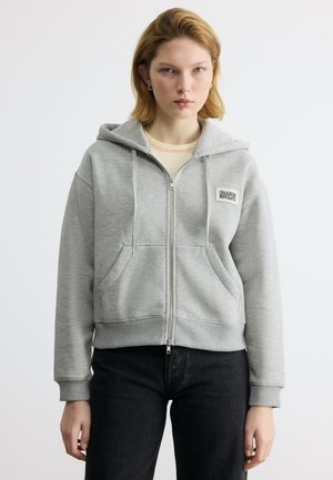 Jonge volwassene die een lichtgrijze hoodie met ritssluiting en zakken draagt, en een zwarte broek, staand tegen een effen witte achtergrond.