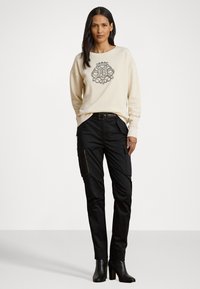 Donna che indossa una felpa color crema con un design del logo intricato, pantaloni cargo neri con tasche e stivaletti neri, in piedi su uno sfondo neutro.