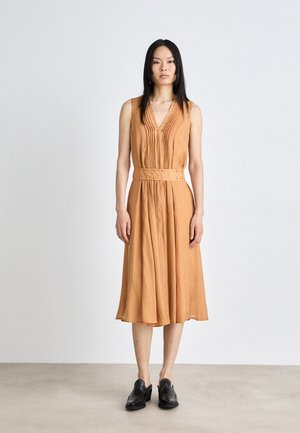 Robe sans manches et décolletée en V en tissu orange chaud avec des plis et une ceinture à la taille arborant des accents. Portée avec des chaussures noires, se tenant sur une surface claire.