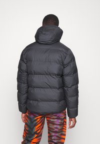 Man som bär en svart vadderad jacka med huva och färgglada mönstrade byxor, stående vänd bortom en enfärgad vit bakgrund.