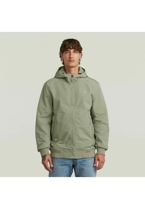 Man draagt een lichtgroene capuchontrui met rits en blauwe spijkerbroek, staand tegen een effen lichte achtergrond.