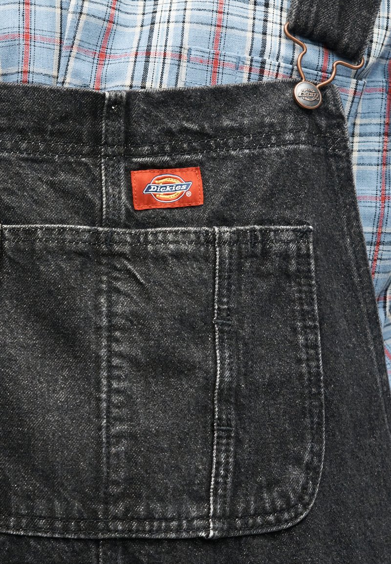 Salopette in denim scuro con una tasca frontale sul petto e etichetta con logo Dickies rossa, indossata sopra una camicia a quadri blu e rossa.