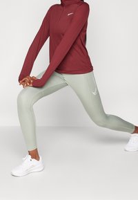 Burgundowa, długorękawowa bluzka sportowa z zamkiem, zestawiona z jasnoszarymi legginsami z delikatnym logo. Białe buty sportowe dopełniają całość.