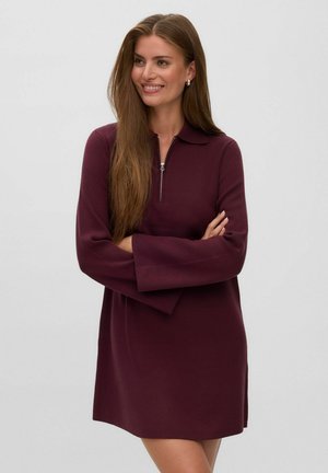 Femme aux longs cheveux bruns portant une robe à manches longues violet foncé avec un col à fermeture éclair, souriante et debout, les bras croisés.