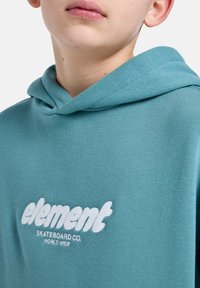 Tyrkysová mikina s kapucí na stažení a měkkou texturou. Na přední straně je umístěno bílé vyšívané logo "element SKATEBOARD CO. WORLD WIDE".