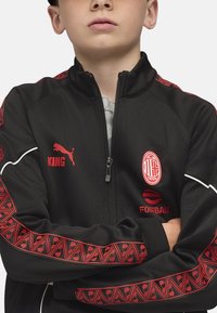 Chaqueta deportiva negra con cremallera frontal, logos rojos y acentos con patrón en las mangas. Presenta el logo de Puma y el texto "KING".
