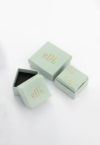 Quadratische, mintfarbene Papierboxen mit einem goldenen Logo, eine davon geöffnet und zeigt ein schwarzes Innenleben, mit glatter Textur und minimalistischem Design.