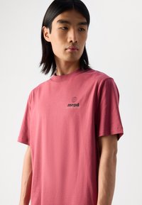 Tricou din bumbac roz, cu mâneci scurte și guler rotund, decorat cu un mic grafic al globului și text pe partea stângă.
