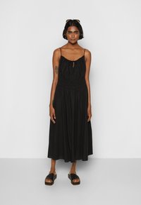 Vestuário preto, sem mangas, com cintura franzida e detalhe de bolsa frontal; usado com sandálias de plataforma pretas. Tecido suave, design até ao meio da coxa.