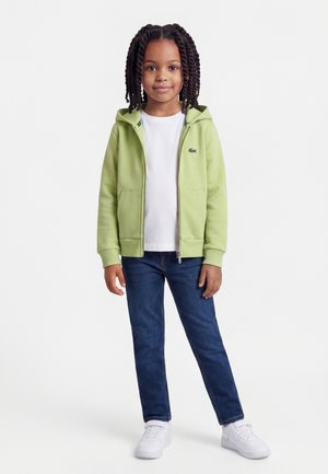 Enfant aux cheveux tressés portant un sweat à capuche zippé vert clair, un t-shirt blanc, un jean bleu et des baskets blanches, debout devant un fond blanc.