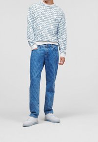 Felpa bianca con stampa blu "LAGERFELD", abbinata a jeans azzurri chiari con un motivo simile. Il modello indossa sneakers bianche.