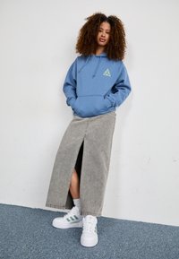 Blauwe hoodie met een voorzak en driehoekig logo, gecombineerd met een lange grijze jeansrok met een split aan de voorkant, en witte sneakers met groene strepen.