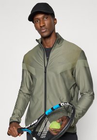 Πράσινο αθλητικό μπουφάν με φερμουάρ μπροστά, στρωματοποιημένο σχέδιο, υφασμάτα σημεία και λογότυπο BOSS. Άτομο κρατά μια μαύρη ρακέτα τένις και μια μπάλα.
