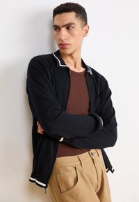 Cardigan nero con zip, colletto e polsini bianchi, texture in maglia, indossato sopra una camicia marrone, abbinato a pantaloni beige. Braccia incrociate contro un muro.