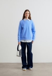 Γυναίκα με μακριά καστανά μαλλιά, φοράει μπλε φούτερ, λευκό πουκάμισο με γιακά, σκούρο παντελόνι, ασημί παπούτσια, κρατώντας μια αντανακλαστική ασημί τσάντα.