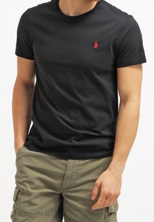 Homme portant un t-shirt noir près du corps avec un petit logo rouge brodé sur la poitrine et un short cargo vert olive avec des poches.