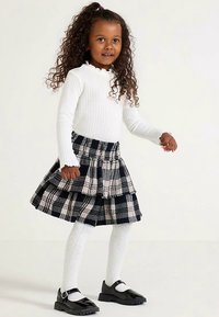 Weißes, geripptes Langarmshirt kombiniert mit einem gestuften karierten Rock in Schwarz und Weiß, ergänzt durch weiße Strumpfhosen und schwarze Lackschuhe.