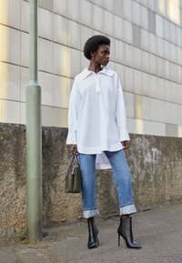 Hvid oversized skjorte med lange ærmer, blå jeans med opkrøllede ben, sorte ankelstøvler og en mørkegrøn taske. Tekstureret væg i baggrunden.
